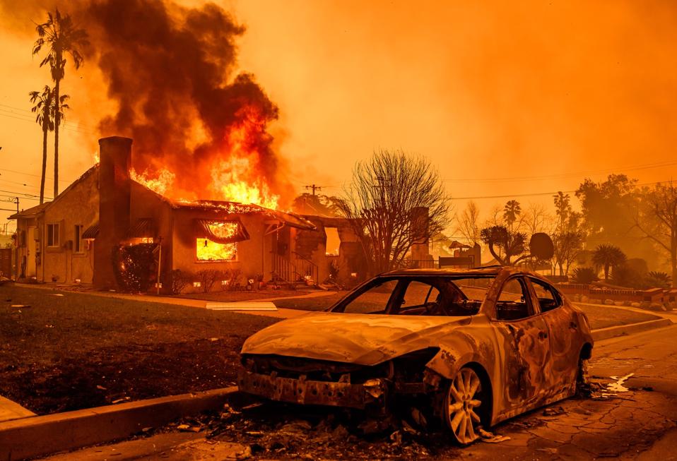 A burned out car in front of a burning house.